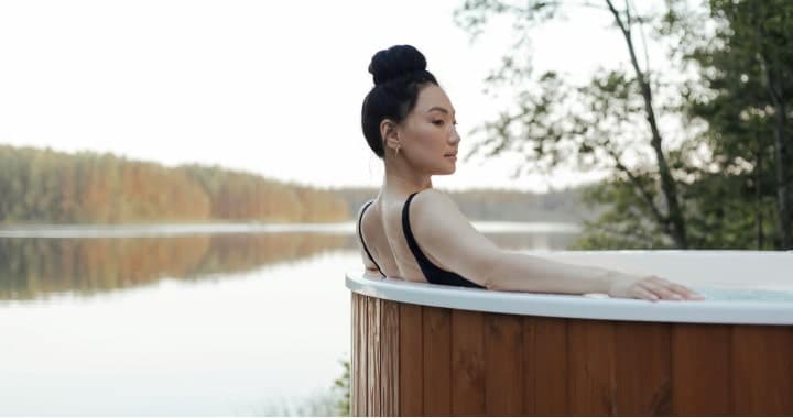 Person relaxing in an outdoor hot tub by a serene lake.